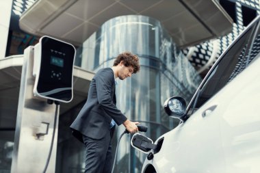 Below view closeup image of progressive black suit businessman recharge battery of his electric vehicle from public charging station. Renewable and alternative energy powered car concept.