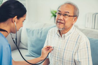 Caring young female doctor examining her contented senior patient with stethoscope in living room. Medical service for elderly, elderly sickness, declining health.