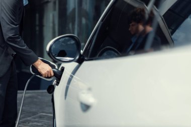 Closeup progressive black suit businessman insert EV charger plug from public charging station to his electric car. Eco friendly electric rechargeable car with zero CO2 emission.