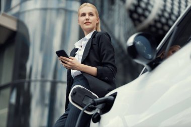 Businesswoman wearing black suit using smartphone, leaning on electric car recharge battery at charging station in city residential building with condos and apartment. Progressive lifestyle concept.