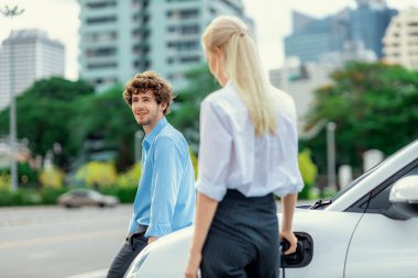 Progressive businessman and businesswoman with electric car parking and connected to public charging station before driving around city center. Eco friendly rechargeable car powered by clean energy.