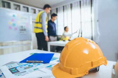 Concept of engineering equipments of protective helmet for competent engineer on piles of paper works. Blurred image of engineers and employer working together at the desk.