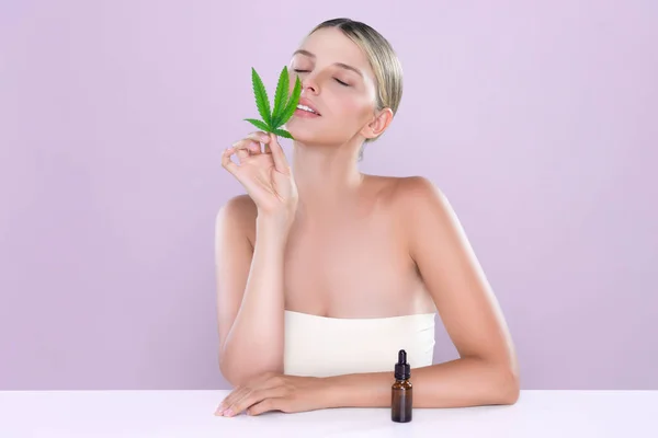 Alluring portrait of beautiful woman in pink isolated background holding green leaf with marijuan extracted bottle for skincare treatment product. Cannabis CBD oil for cosmetology and beauty concept.