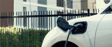 Kamu şarj istasyonunda EV şarj cihazına bağlı yakın çekim elektrikli araç yenilenebilir temiz enerji ile destekleniyor ve arka planda konut inşaatı yenilikçi kavram olarak kullanılıyor.