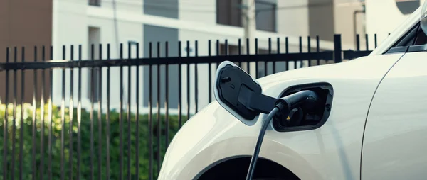Closeup electric vehicle plugged in with EV charger at public charging station powered by renewable clean energy with residential building in background as progressive concept.