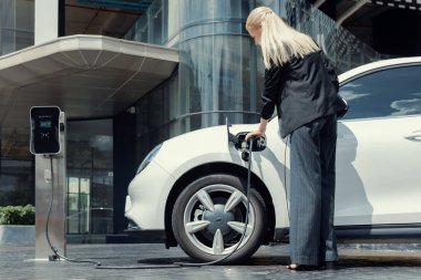 Progressive businesswoman insert charger plug from charging station to her electric vehicle with apartment condo building in background. Eco friendly rechargeable car powered by sustainable energy.