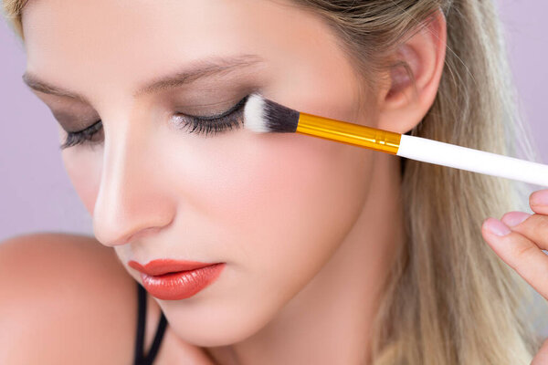 Крупный план красивая девушка с безупречным нанесением заманчивой тени для глаз макияж с кистью подводки для глаз. Процесс косметической живописи лица на красивой молодой женщине с идеальной чистой кожей на розовом изолированном фоне