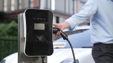 İlerici işadamı, şehir merkezinde dolaşmadan önce şarj cihazından elektrikli arabasına şarj prizi takıyor. Çevre dostu şarj edilebilir araba, sürdürülebilir ve temiz enerjiyle çalışıyor..