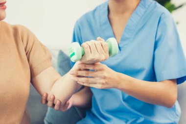 Caregiver helping contented senior woman exercise with dumbbell at home. Rehab for elder, senior medical care.