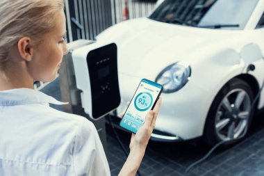 Suit-clad progressive businesswoman look at EV cars battery status from her phone while standing on a charging station with a power cable plug and a renewable energy-powered electric vehicle.