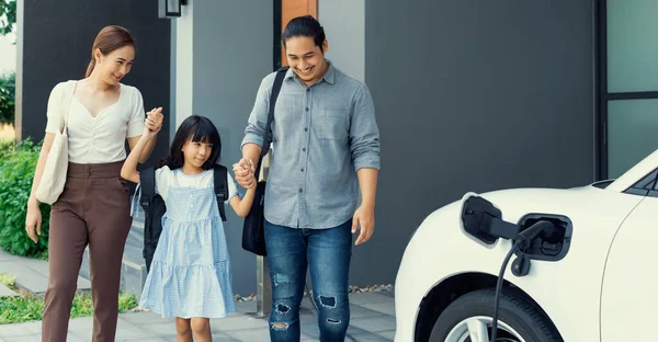 Progressive young parents and daughter with electric vehicle and home charging station. Green and clean energy from electric vehicles for healthy environment. Eco power from renewable source at home.