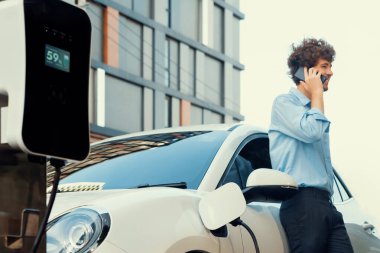 Progressive businessman talking on the phone, leaning on electric car recharging with public EV charging station, apartment condo residential building on the background as green city lifestyle.