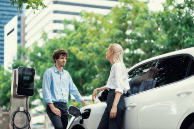 Progressive businessman and businesswoman with electric car parking and connected to public charging station before driving around city center. Eco friendly rechargeable car powered by clean energy.