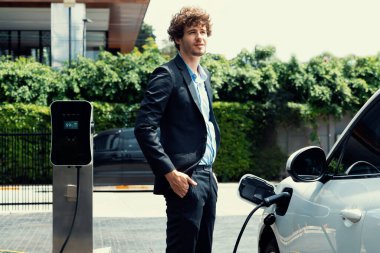 Progressive businessman wearing black suit with electric car recharging at public parking car charging station at modern city residential area. Eco friendly rechargeable car powered by clean energy.