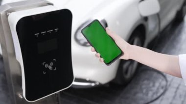 Taklit uzay için şarj istasyonuna bağlı elektrikli aracın yeşil ekran enerji durumunu gösteren bir model telefon. Temiz çevre için ilerici bir kavram. EV, yeşil yenilenebilir enerjiden güç alıyor.