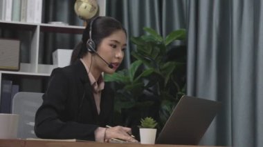 Kulaklık ve mikrofon takan iş kadını masasında çalışıyor ve dizüstü bilgisayar kullanıyor. Uzaktan kumandalı online toplantı için dizüstü bilgisayar kullanan hevesli kadın çalışanlar, ofiste iş videosu konferansı.