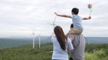 İlerici mutlu aile kavramı rüzgâr türbini çiftliğinde eğleniyor. Rüzgar türbini jeneratörüyle gelen elektrik jeneratörü kırsal bölgede tepe ve ufukta dağ var..