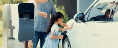 Closeup focus hand insert EV charger plug to electric vehicle at home charging station with blurred family in background. Progressive concept of green and clean energy to reduce CO2 emission by EV car