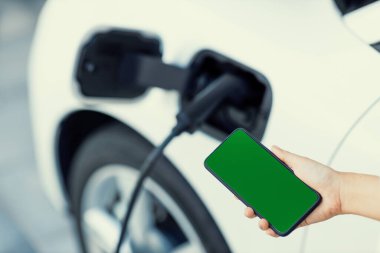 Taklit uzay için şarj istasyonuna bağlı elektrikli aracın yeşil ekran enerji durumunu gösteren bir model telefon. Temiz çevre için ilerici bir kavram. EV, yeşil yenilenebilir enerjiden güç alıyor.