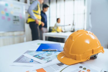 Concept of engineering equipments of protective helmet for competent engineer on piles of paper works. Blurred image of engineers and employer working together at the desk.