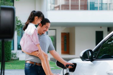 Progressive father and daughter returned from school in electric vehicle that is being charged at home. Electric vehicle driven by renewable clean energy. Home charging station concept for environment