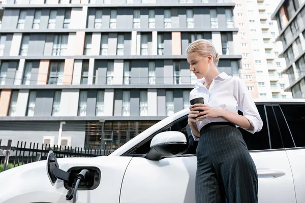 Businesswoman Drinking Coffee Leaning Electric Vehicle Recharging Public Charging Station — Stockfoto