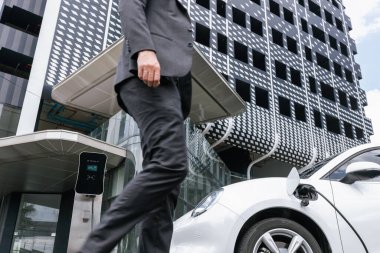 Closeup progressive businessman with electric car recharging at public charging station with background of city residential building. Eco friendly car powered by alternative clean energy.