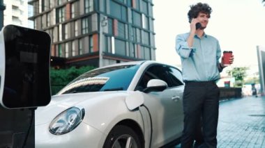Takım elbiseli, hırslı bir işadamı elektrik kablosu ve yenilenebilir enerji enerjili bir elektrikli araçla şarj istasyonunda dururken elektrikli aracına yaslanıyor..