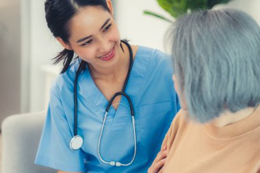A caregiver rest her hands on the shoulders of a contented senior patient while she sitting on the sofa at home.