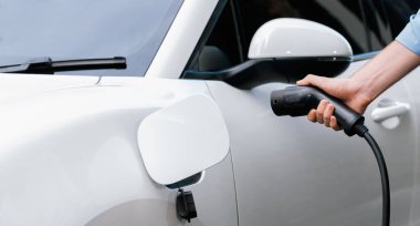 Closeup progressive man holding EV charger plug from public charging station for electric vehicle with background of residential building as concept eco-friendly sustainability energy car concept.