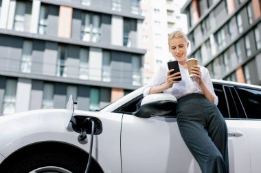 Focus businessman using phone, leaning on electric vehicle, holding coffee with blurred city residential condo buildings in background as progressive lifestyle by renewable and sustainable EV car.
