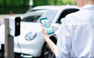 Suit-clad progressive businesswoman look at EV cars battery status from her phone while standing on a charging station with a power cable plug and a renewable energy-powered electric vehicle.