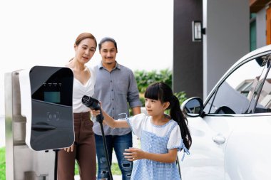 Progressive young parent teach daughter how to recharge or refuel EV car at home charging station. Green and clean energy from electric vehicle for healthy environment. Eco power from renewable source