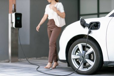 Focus image of electric vehicle recharging battery at home charging station with blurred woman walking in the background. Progressive concept of green energy technology applied in daily lifestyle.