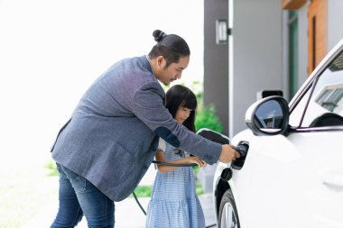 Progressive father and daughter plugs EV charger from home charging station to electric vehicle. Future eco-friendly car with EV cars powered by renewable source of clean energy.