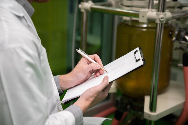 Closeup apothecary scientist using a clipboard and pen to record information from a CBD oil extractor and a scientific machine used to create medicinal cannabis products.