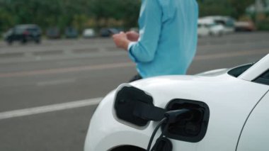 Takım elbiseli, hırslı bir işadamı elektrik kablosu ve yenilenebilir enerji enerjili bir elektrikli araçla şarj istasyonunda dururken elektrikli aracına yaslanıyor..