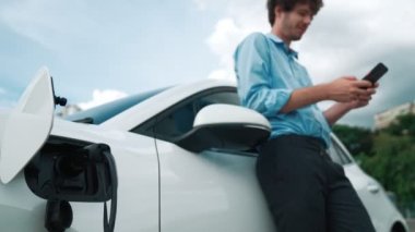 Takım elbiseli, hırslı bir işadamı elektrik kablosu ve yenilenebilir enerji enerjili bir elektrikli araçla şarj istasyonunda dururken elektrikli aracına yaslanıyor..