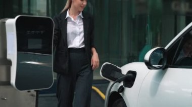 İlerici iş kadını, şehir merkezinde dolaşmadan önce şarj cihazının fişini şarj istasyonundan elektrikli arabasına çekti. Çevre dostu şarj edilebilir araba, sürdürülebilir ve temiz enerjiyle çalışıyor..
