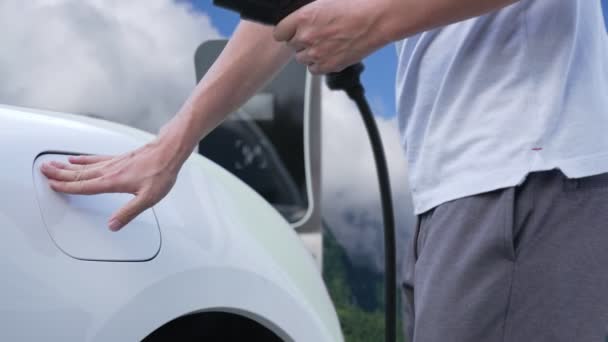 充電ポイントで電気自動車を充電する進歩的な男 きれいな環境のためのクリーンで再生可能なエネルギーを搭載したEvの自然と山を旅 — ストック動画