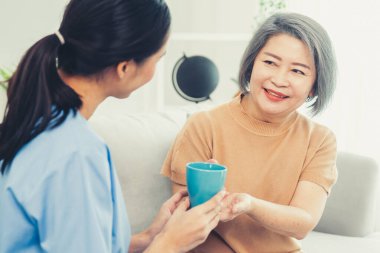 Female care taker serving her contented senior patient with a cup of coffee at home, smiling to each other. Medical care for pensioners, Home health care service.