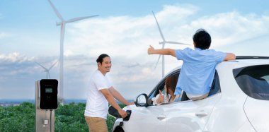 İlerici mutlu aile kavramı elektrikli araçla rüzgar çiftliğinde eğleniyor. Rüzgar türbini jeneratöründen gelen temiz yenilenebilir enerji ile şarj edilen elektrikli araç..