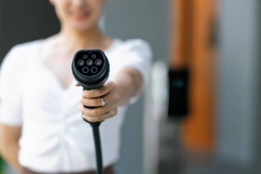 Focus hand holding and pointing EV charger plug for electric vehicle at camera with blur woman in background from home charging station as progressive idea of alternative energy for ecology concern.