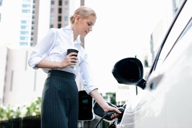 Businesswoman drinking coffee, leaning on electric vehicle recharging at public charging station with residential apartment condos building in background as progressive lifestyle by eco-friendly car.