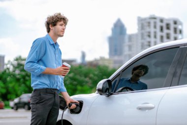 Progressive eco-friendly concept of parking EV car at public electric-powered charging station in city with blur background of businessman leaning on recharging-electric vehicle with coffee.