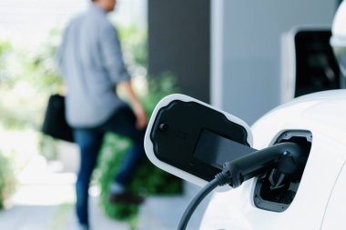 Focus electric car charging at home charging station with blurred progressive man walking in the background. Electric car using renewable clean for eco-friendly concept.