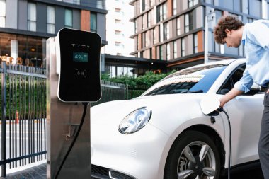 Progressive businessman insert charger plug from charging station to his electric vehicle with apartment condo building in background. Eco friendly rechargeable car powered by sustainable energy.