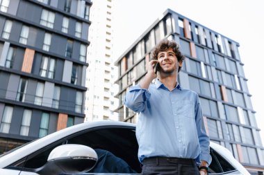 Progressive businessman talking on the phone, leaning on electric car recharging with public EV charging station, apartment condo residential building on the background as green city lifestyle.