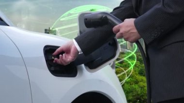 İlerleyen yeşil şehir ESG sembolü, elektrikli araçla birlikte. İş adamı EV arabalarını şarj etmek için şarj aletini şarj ediyor. Şehirdeki şarj istasyonunda ekolojik ve yeşil semboller var..