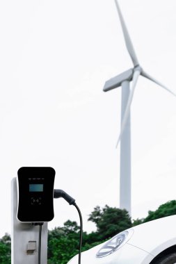Progressive combination of wind turbine and EV car, future energy infrastructure. Electric vehicle being charged at charging station powered by renewable energy from wind turbine in the countryside.